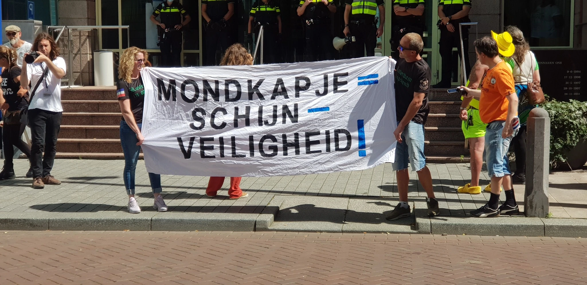 Demonstranten houden een groot wit spandoek vast met de tekst “Mondkapje = schijnveiligheid”, terwijl politieagenten op de achtergrond toekijken.